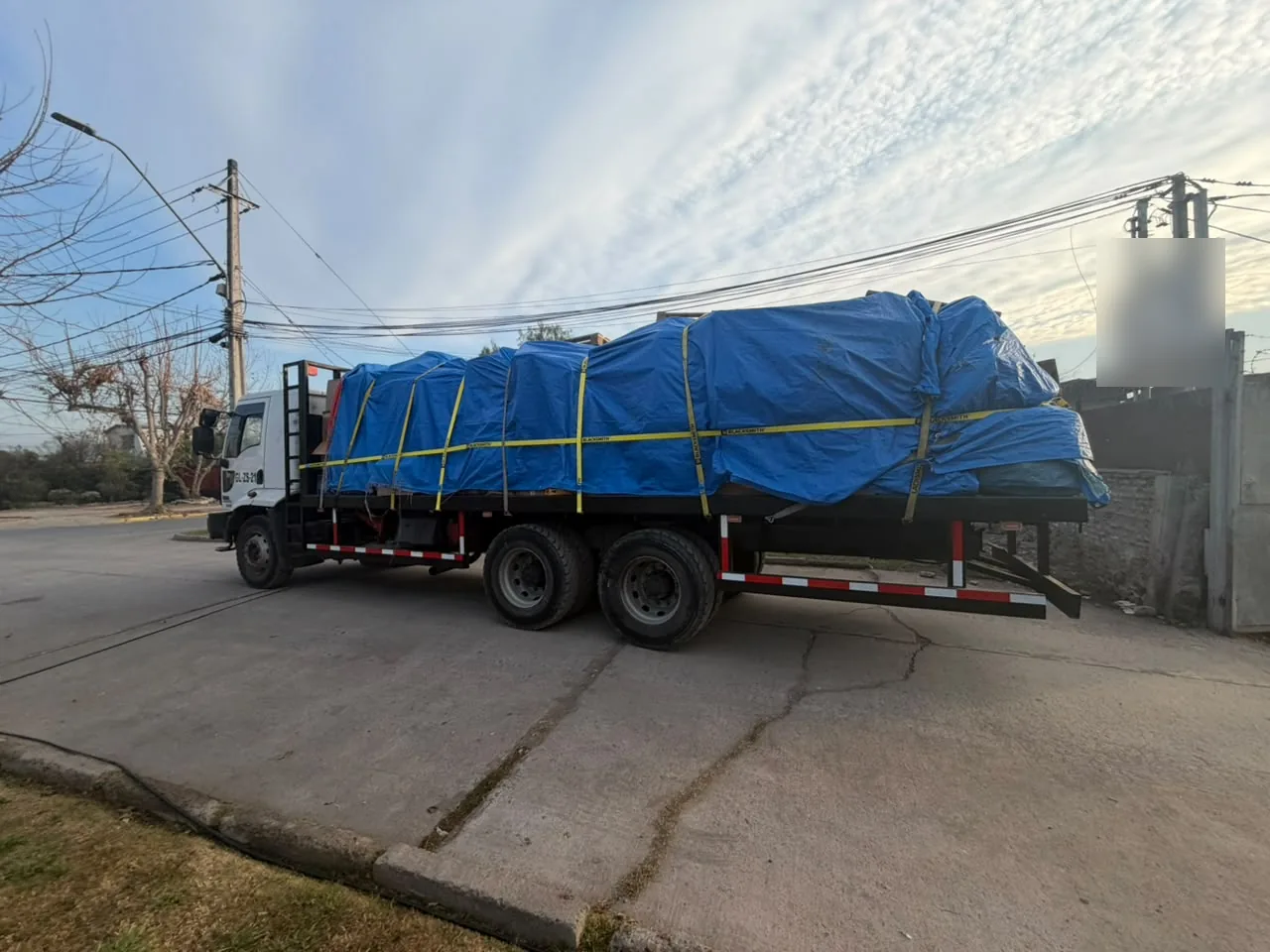 Camión plano con carga cubierta con lona azul y correas amarillas, estacionado en una calle