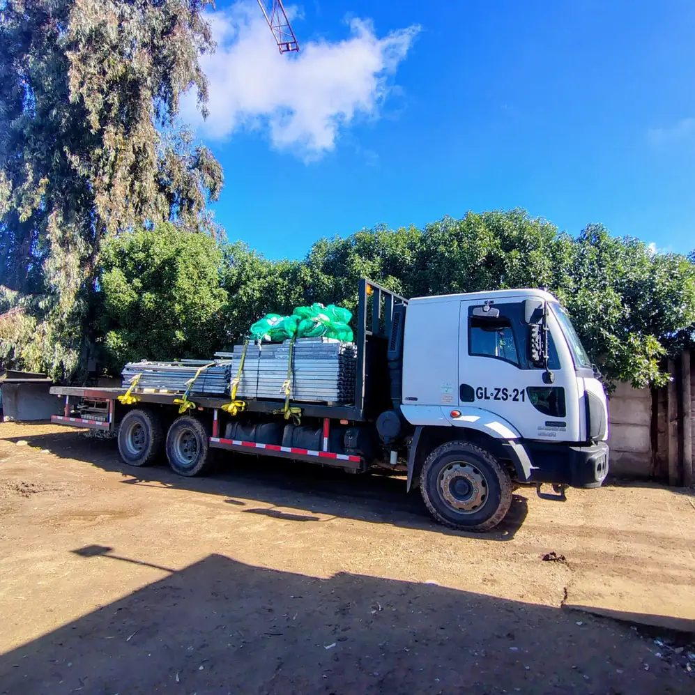 Camión plano con estructuras metálicas apiladas y una malla verde, amarradas con correas, en un patio