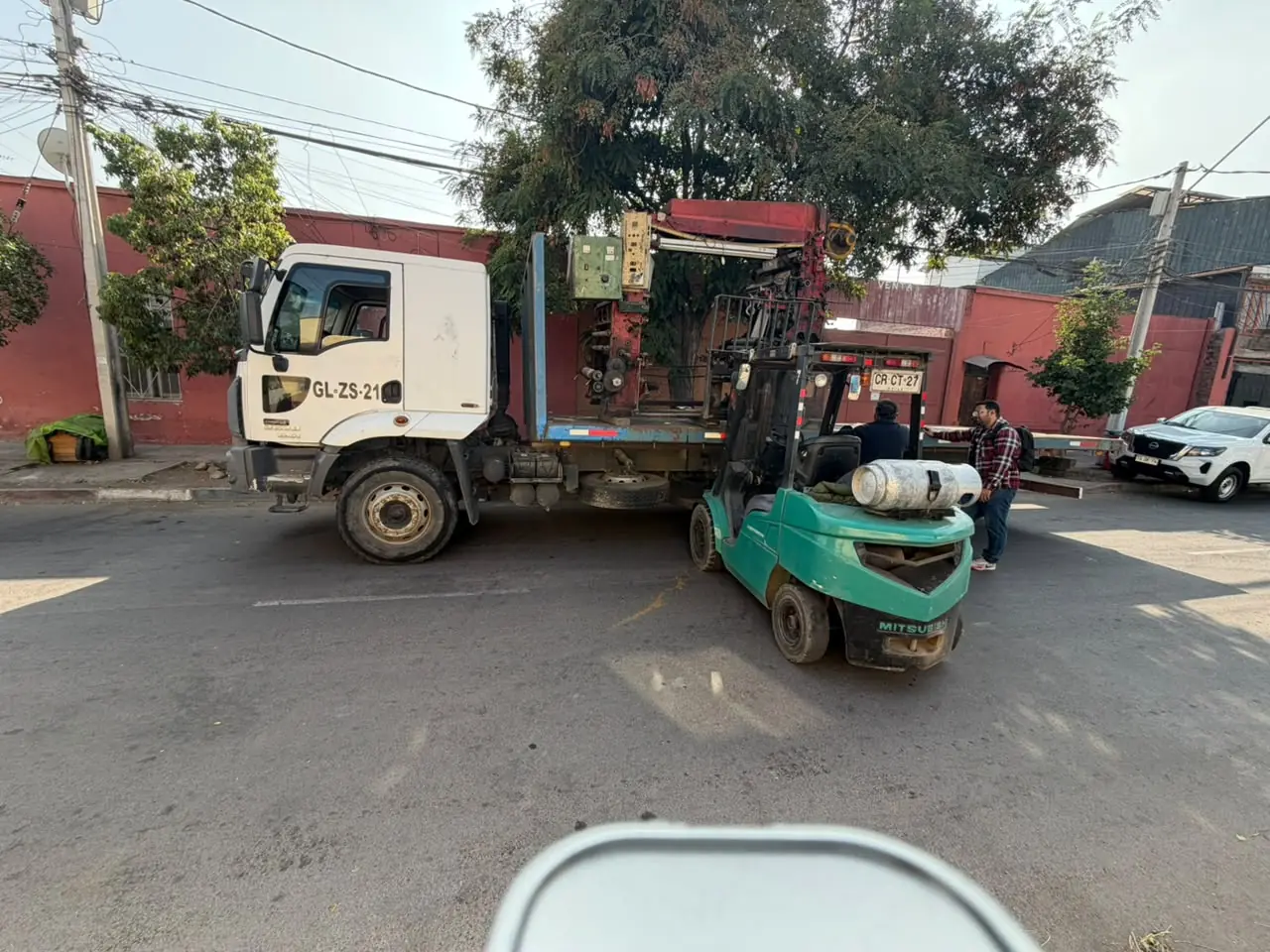 Camión plano con una máquina industrial sobre la plataforma y un montacargas verde junto a la parte trasera, con dos personas, en una calle