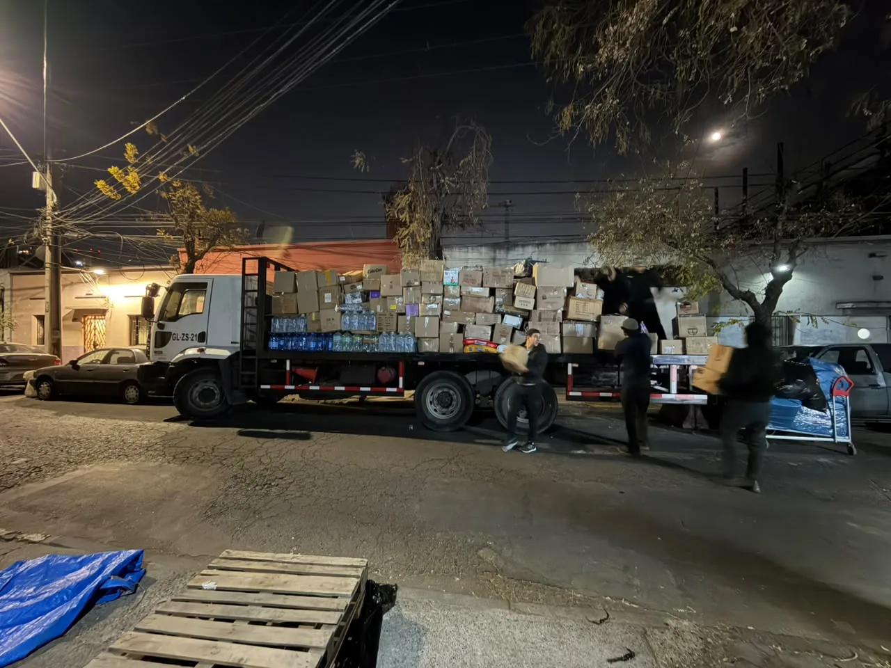 Camión plano cargado con cajas de cartón y bidones de agua mientras varias personas descargan de noche