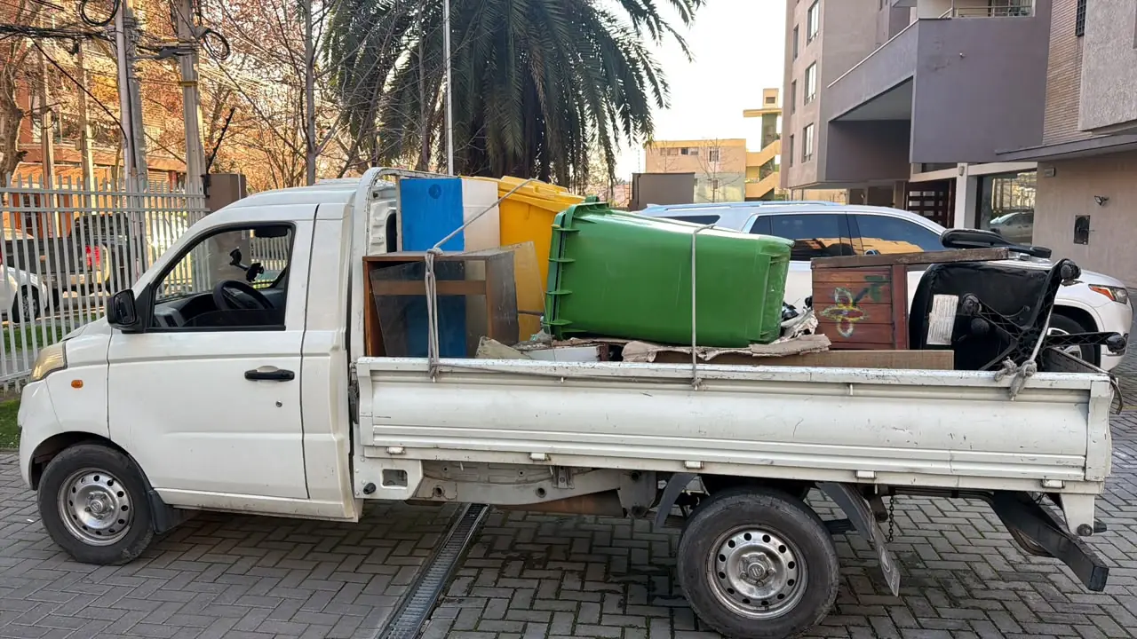 Camioneta blanca de plataforma cargada con muebles viejos, un contenedor de basura verde y tablas, amarrados con cuerdas