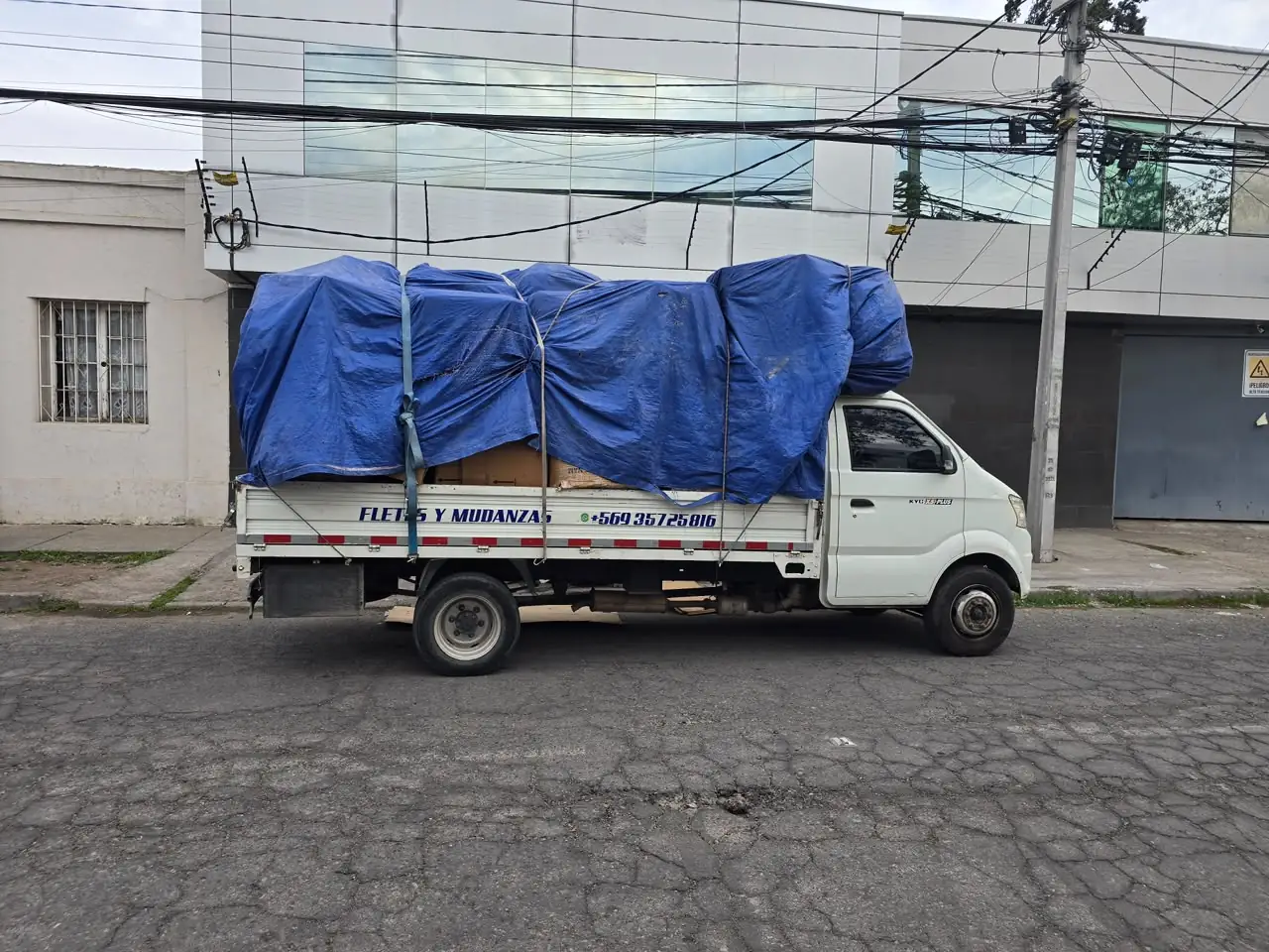 Camioneta KYC X5 Plus con la carga cubierta con una lona azul y amarrada con correas; en la baranda dice «Fletes y Mudanzas»