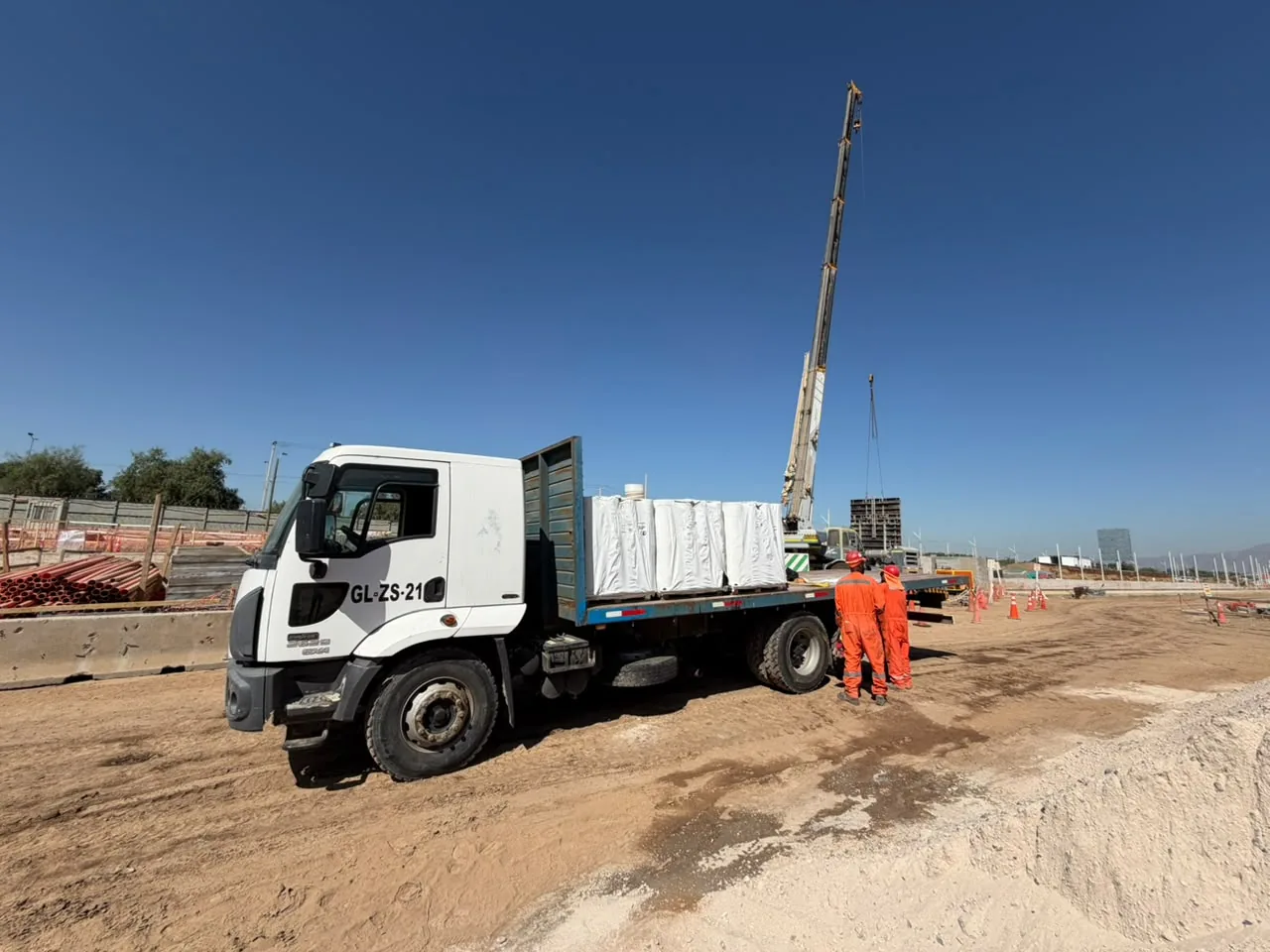 Camión plano con bultos blancos sobre pallets en una obra, con una grúa, tubos naranjos y dos trabajadores al fondo