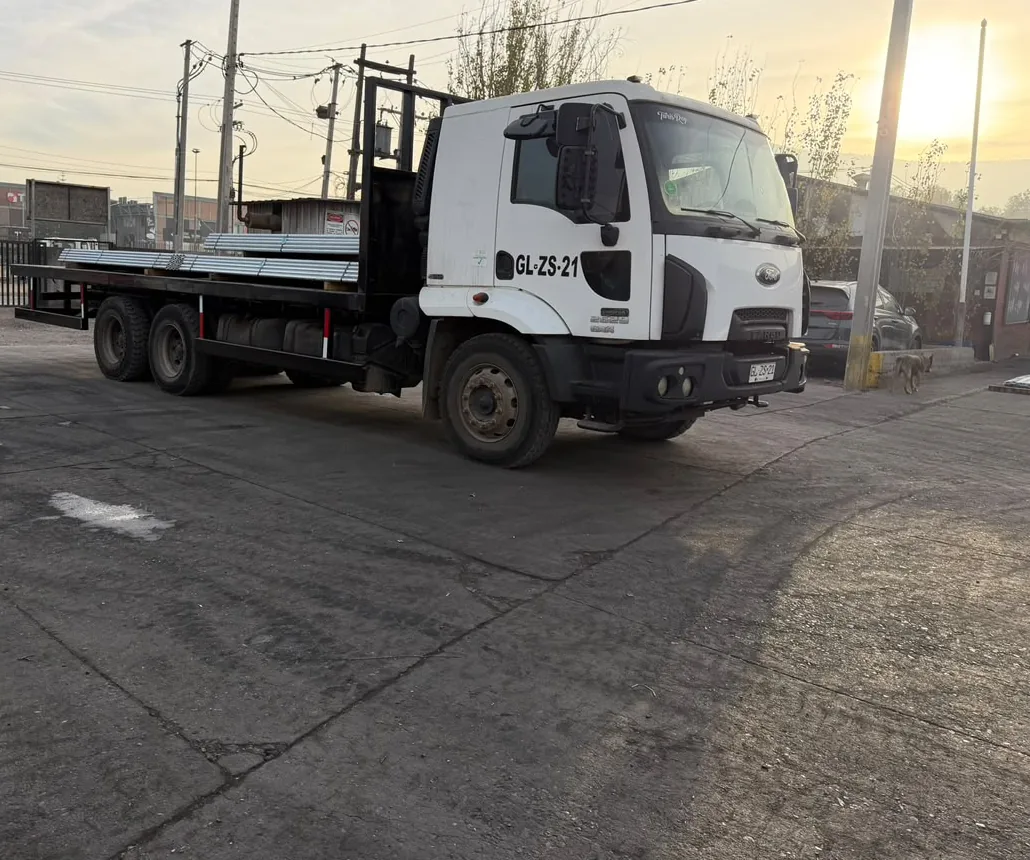 Camión plano con tubos metálicos sobre la plataforma, estacionado en un patio de cemento